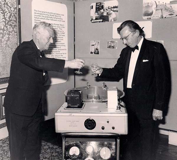 Two men in suits holding glasses of wine over the bowl of a Travenol open-bowl dialysis machine (twin coil)